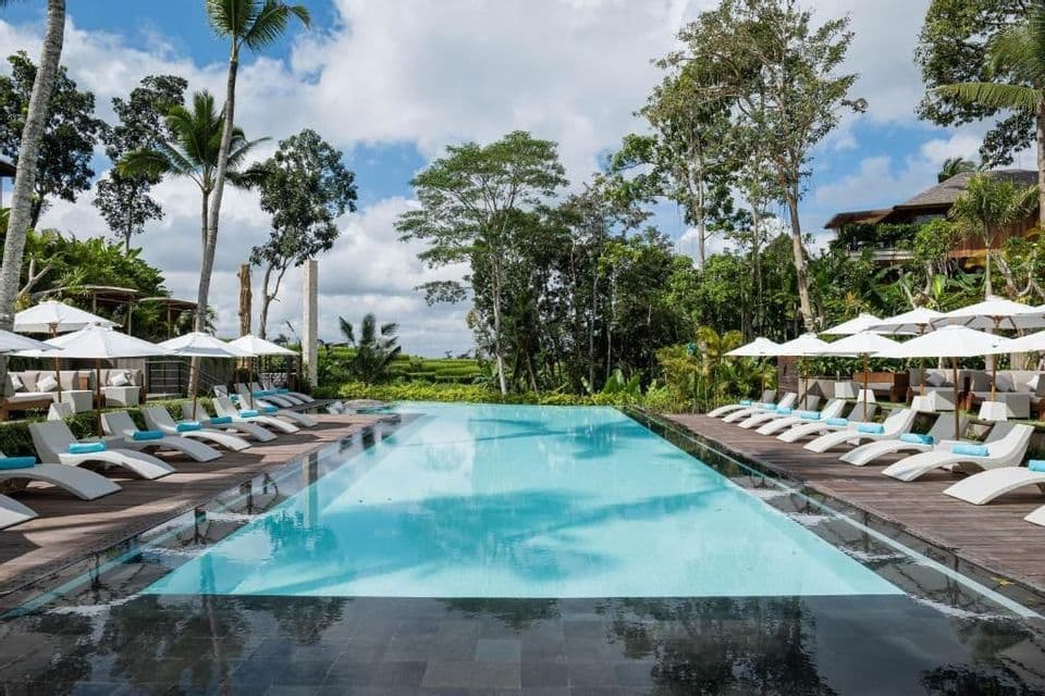 Une longue piscine dans un complexe tropical, flanquée de rangées de chaises longues blanches et de parasols sous un ciel partiellement nuageux.