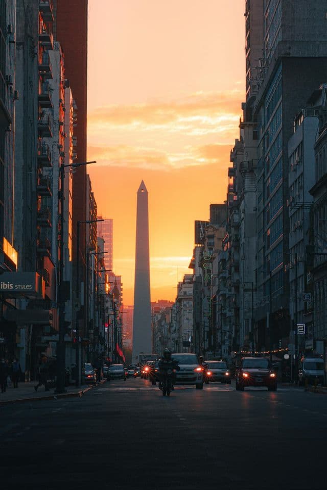 Un alto obelisco se alza al final de una calle de la ciudad flanqueada por edificios, con tráfico en la carretera durante un cálido atardecer.