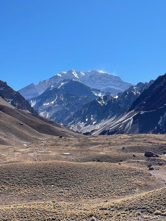 Un vasto valle seco con vegetación escasa conduce hacia una sierra maciza y nevada bajo un cielo azul claro.