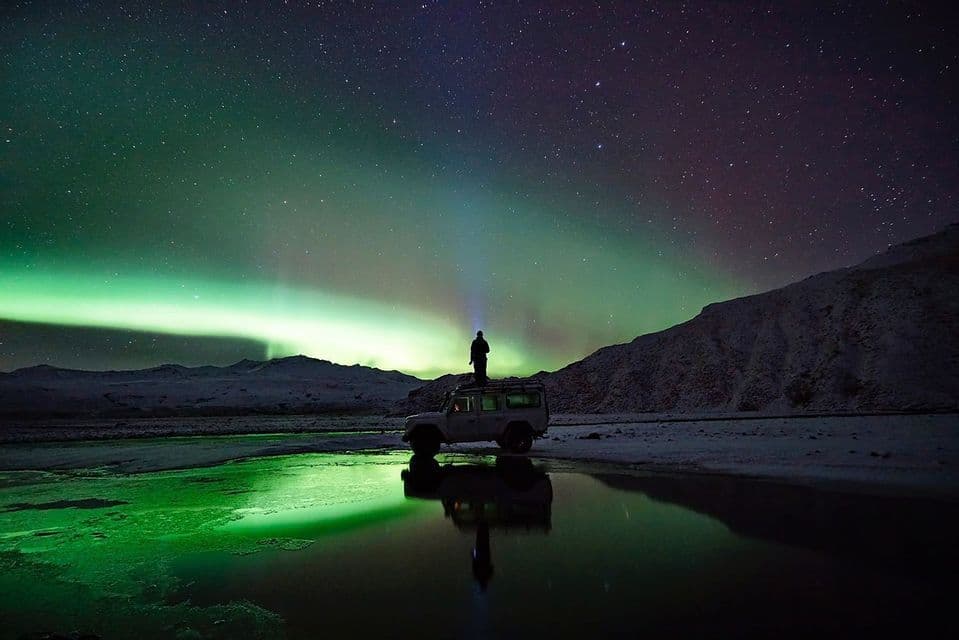 A person stands on an SUV under a starry sky with the green aurora borealis, which is reflected in a calm body of water.