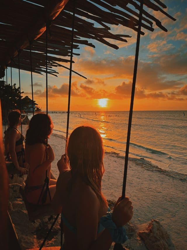 Un gruppo WeRoad siede su delle altalene sotto un tetto di paglia, ammirando il tramonto sull'oceano.