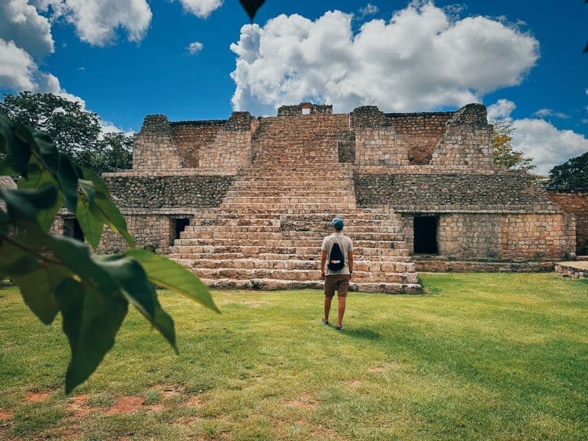 Un uomo visto di spalle, in piedi su un prato verde e che guarda una grande antica piramide di pietra sotto un cielo blu con nuvole.