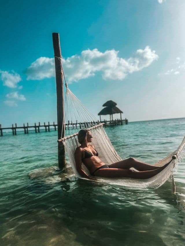 Una mujer en bikini negro se relaja en una hamaca blanca suspendida sobre el agua turquesa del océano con un muelle al fondo.