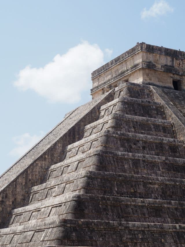 L'angolo di una grande piramide a gradoni di pietra con ripide scale, vista dal basso contro un cielo azzurro e una nuvola bianca.
