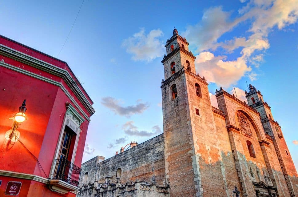 Una vista en contrapicado de una gran catedral de piedra iluminada por el atardecer, junto a un vibrante edificio rojo bajo un cielo parcialmente nublado.