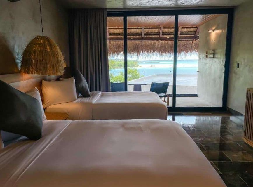 Deux lits avec des draps blancs dans une chambre d'hôtel rustique avec une grande porte vitrée donnant sur un balcon avec vue sur la mer.