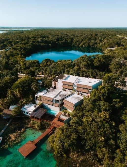 Vue aérienne d'un complexe moderne avec une jetée sur un lagon turquoise, entouré d'une jungle luxuriante et d'un lac proche.