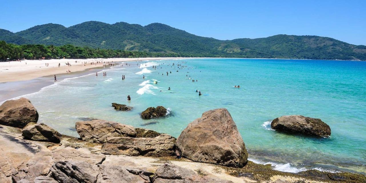 Una spiaggia affollata in una giornata di sole con persone che nuotano in acque turchesi, con montagne verdi e lussureggianti sullo sfondo.