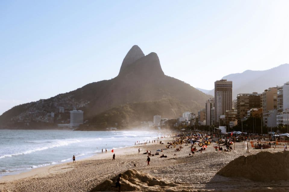 Un'ampia veduta di una spiaggia sabbiosa affollata vicino a una città, con grandi montagne verdi che si ergono sullo sfondo sotto un cielo fosco.