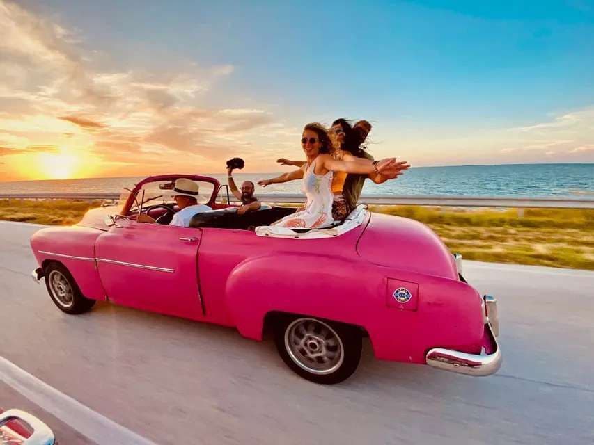 Eine WeRoad-Gruppenreise fährt in einem pinken Cabrio entlang einer Küstenstraße während eines farbenprächtigen Sonnenuntergangs.