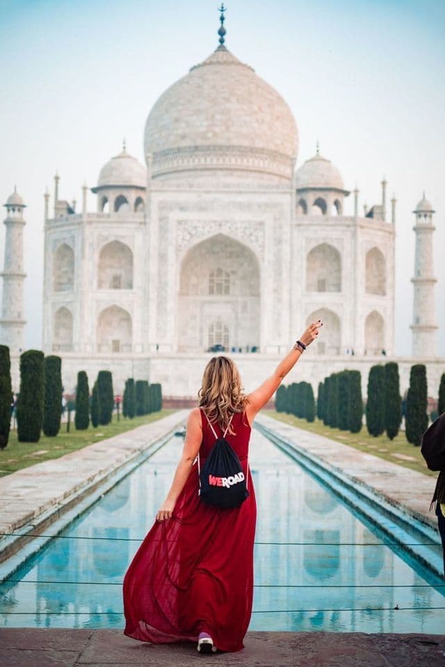 Une femme de dos, vêtue d'une longue robe rouge et portant un sac à dos WeRoad, lève le bras devant le Taj Mahal et son bassin réfléchissant.
