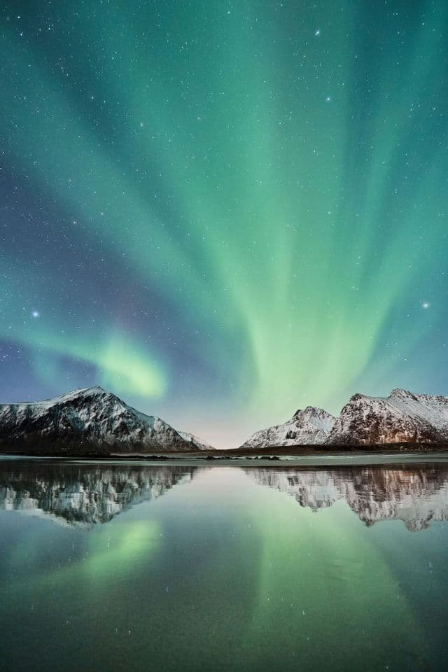 La aurora boreal verde brilla sobre montañas nevadas, reflejándose toda la escena en un cuerpo de agua tranquilo.