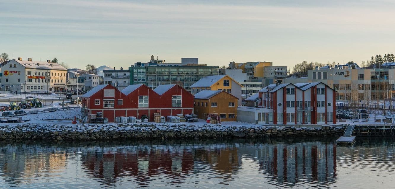 Edifici colorati con tetti innevati si affacciano su un lungomare, le loro riflessioni visibili nell'acqua calma.
