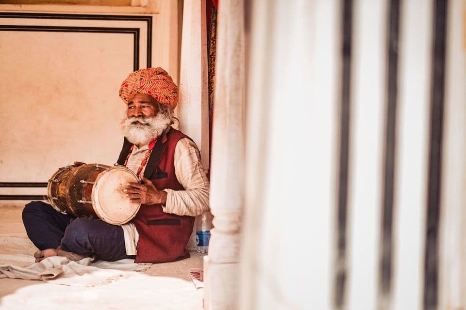 Un uomo con una lunga barba bianca e un turbante rosso siede a gambe incrociate sul pavimento, tenendo un tamburo tradizionale.