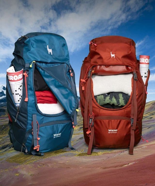 Due zaini da viaggio WeRoad, uno blu e uno color ruggine, pieni di vestiti, affiancati in un paesaggio montano colorato.