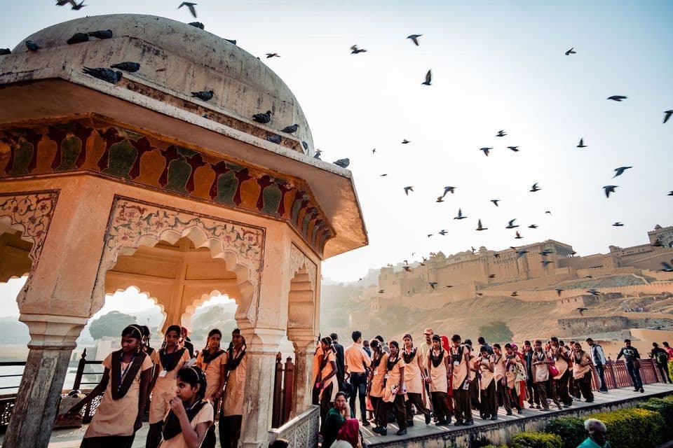 Un gruppo di studentesse in uniforme passa davanti a un padiglione decorato, con uccelli che volano in alto e un grande forte sullo sfondo.