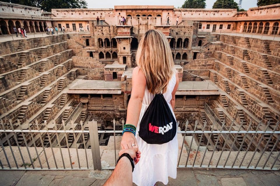 Una donna con uno zaino WeRoad tiene per mano, osservando un grande e antico pozzo a gradini in pietra con scalinate simmetriche.