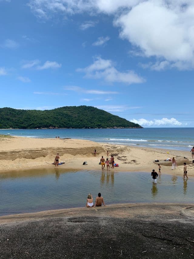 Des gens se relaxant sur une plage de sable avec une piscine naturelle, une colline verdoyante et l'océan en arrière-plan.
