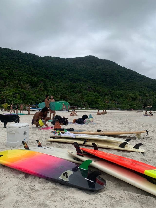 Tablas de surf coloridas en una playa de arena con un grupo de viaje WeRoad preparando su equipo, con una gran colina verde de fondo.