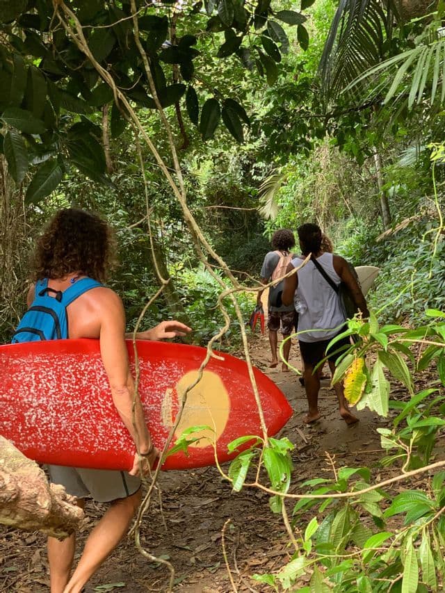 Un viaje en grupo de WeRoad caminando en fila india con tablas de surf por un estrecho sendero de tierra a través de una exuberante selva verde.
