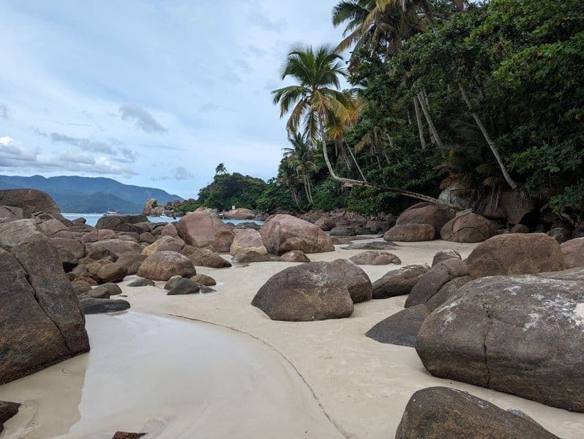 Grandes rocas redondeadas reposan en una playa de arena junto a un bosque tropical con palmeras, con montañas distantes cruzando el agua.