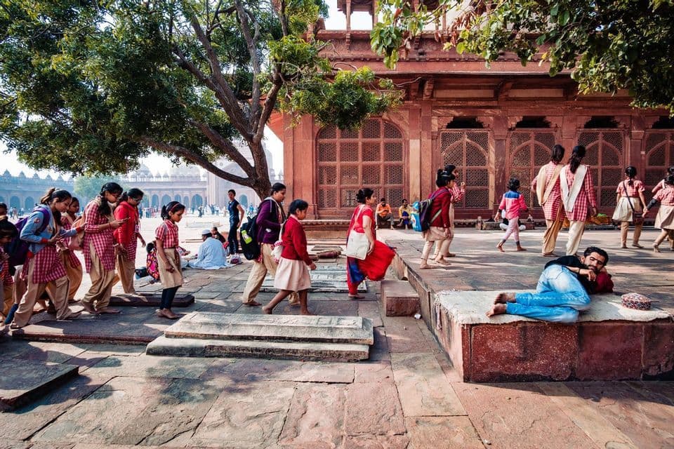 Un gruppo di scolare in divisa attraversa un cortile in pietra, passando accanto a un uomo che riposa su una pedana.