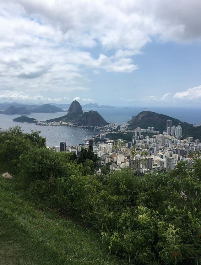 Vue plongeante d'une colline verdoyante et luxuriante surplombant une ville côtière, une baie et un imposant mont conique sous un ciel nuageux.
