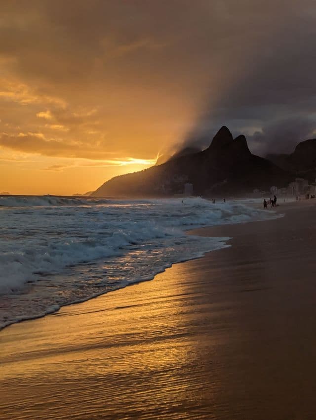 Goldenes Abendlicht spiegelt sich auf nassem Sand, während Wellen ans Ufer schlagen und sich Bergsilhouetten vor einem bewölkten Himmel abzeichnen.