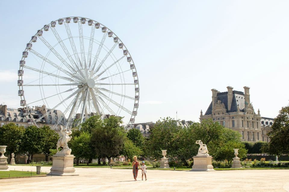 Un adulto e un bambino camminano attraverso un grande parco con statue, una grande ruota panoramica ed edifici storici sotto un cielo sereno.