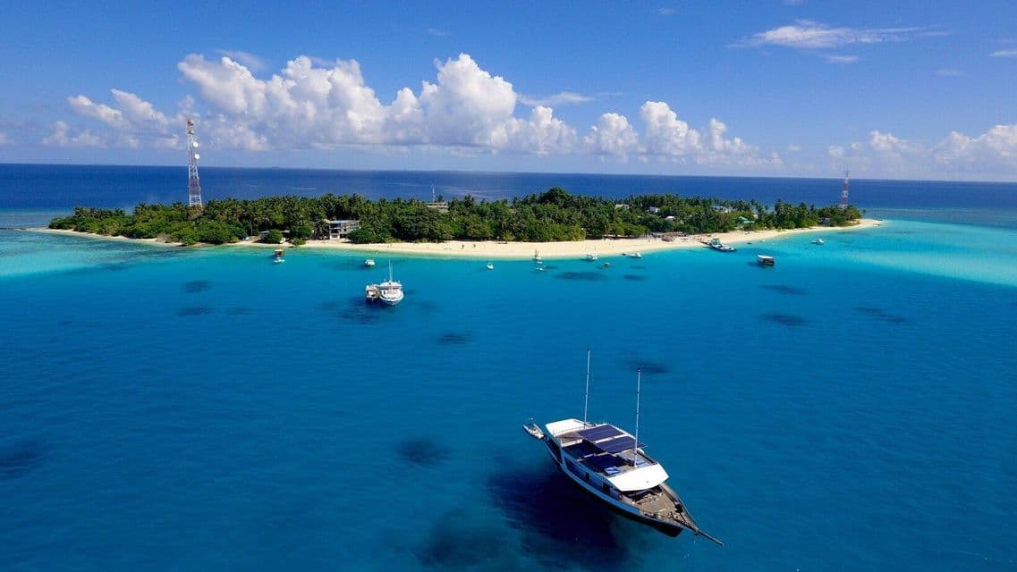 Aerial view of a tropical island with a white sand beach, surrounded by turquoise water and several anchored boats.