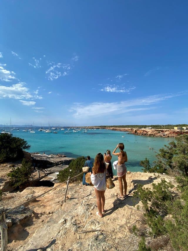 Un viaje en grupo de WeRoad se alza sobre un acantilado rocoso con vistas a una bahía turquesa llena de veleros bajo un cielo azul.