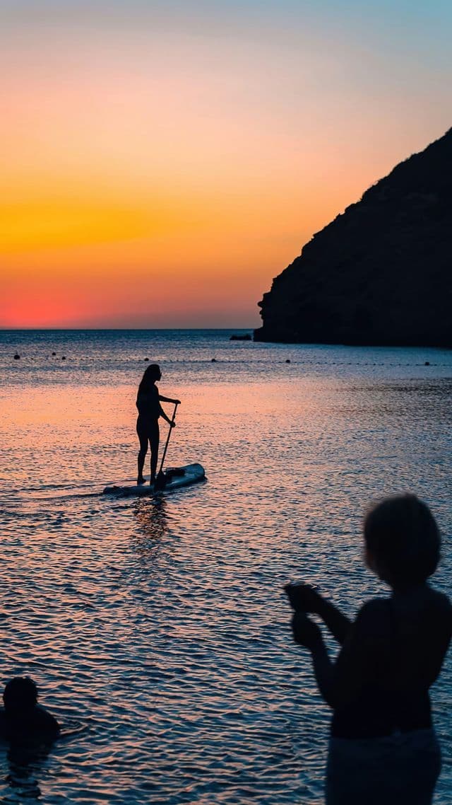 Una persona en una tabla de paddle surf se recorta contra una vibrante puesta de sol naranja sobre el agua.