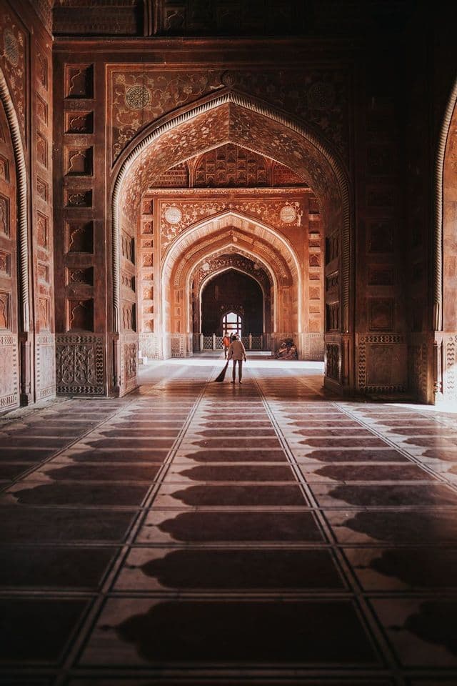 Una persona con una scopa spazza il pavimento piastrellato di un lungo corridoio fiancheggiato da archi in pietra rossa intagliata e ornata che creano lunghe ombre.