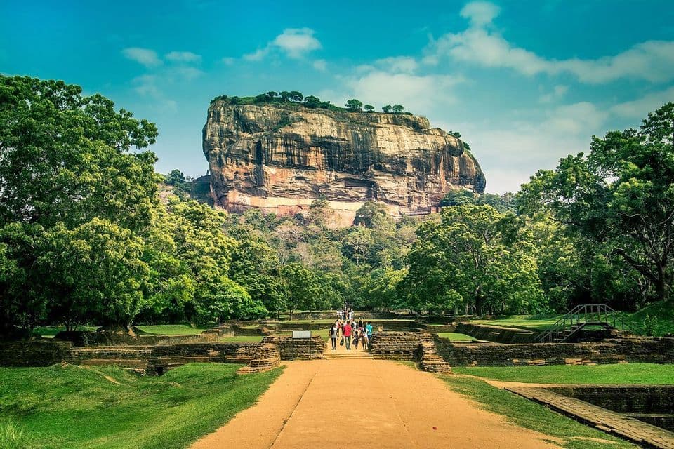 A WeRoad group trip walks on a path through grassy ruins towards a large rock fortress surrounded by lush green trees.