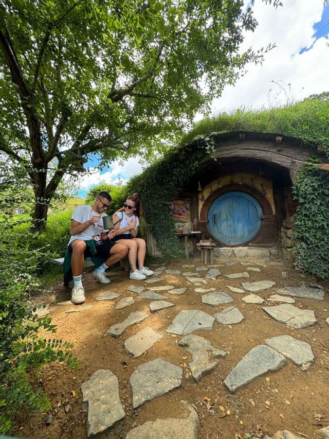 Due persone di un viaggio di gruppo WeRoad siedono su una panchina, sorridendo, fuori da una casa in stile hobbit con una porta blu rotonda.