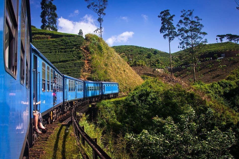 A blue train carrying a WeRoad group trip travels on a curving track through green, terraced hills under a bright blue sky.