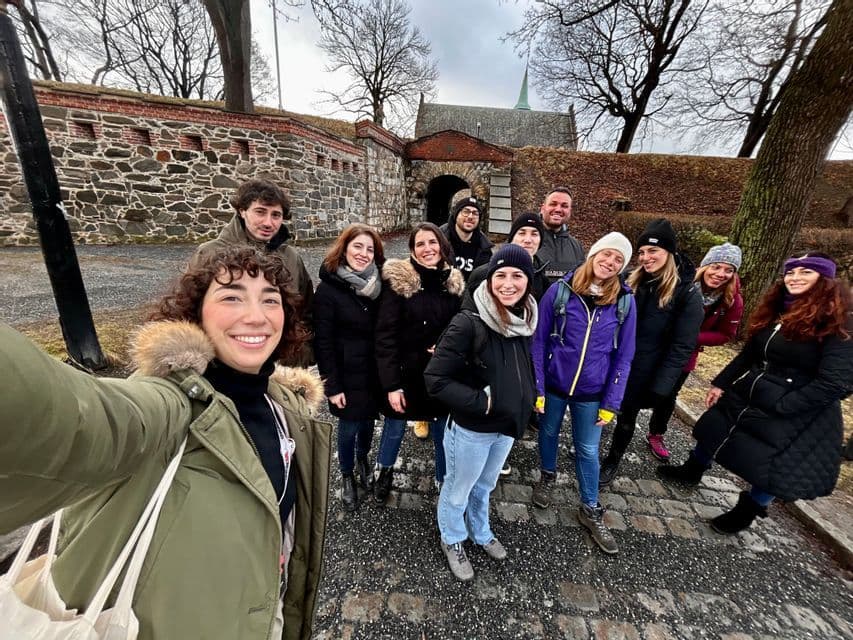 Un gruppo WeRoad in viaggio si scatta un selfie su un sentiero acciottolato davanti a una fortezza storica in pietra, in una giornata nuvolosa.
