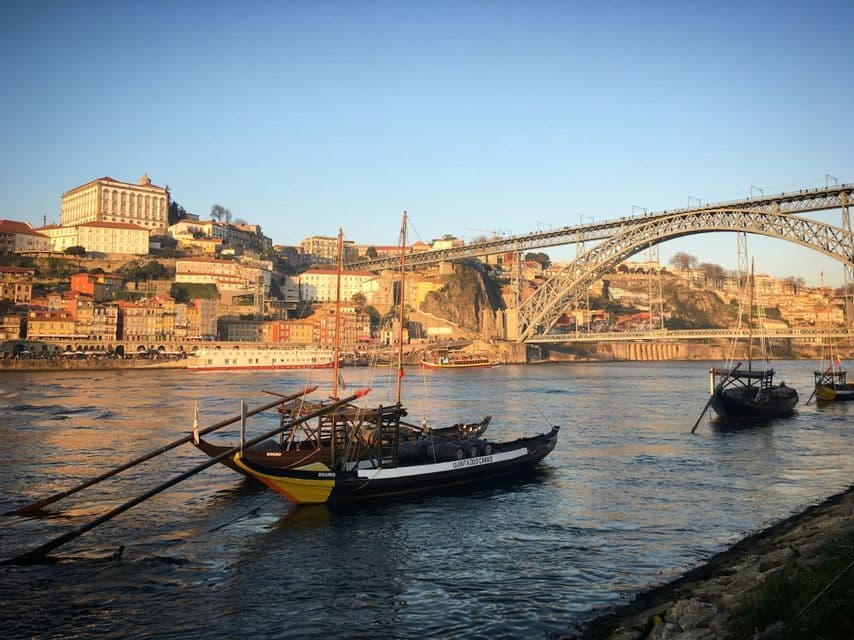 Barche tradizionali ormeggiate su un fiume con una città collinare e un grande ponte metallico ad arco sullo sfondo al tramonto.