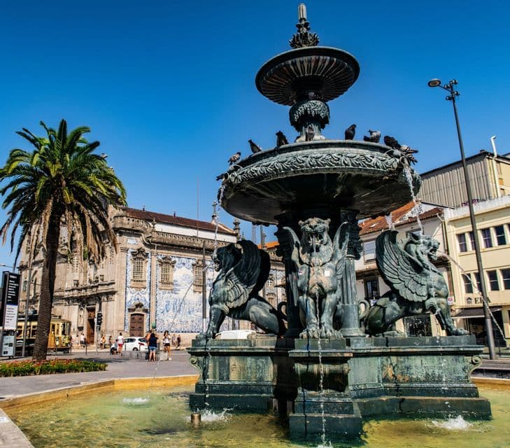 Una grande fontana riccamente decorata con statue di leoni alati in una piazza soleggiata, con una palma e un edificio dalle piastrelle blu sullo sfondo.
