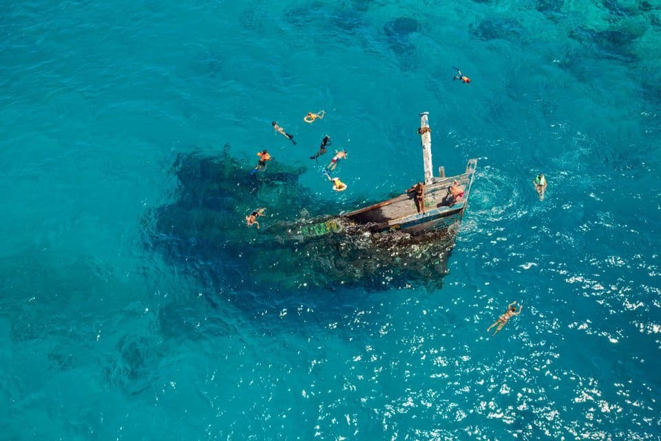 Vue aérienne d'un groupe WeRoad en voyage, faisant de la plongée avec tuba autour d'une épave dans une eau turquoise éclatante.