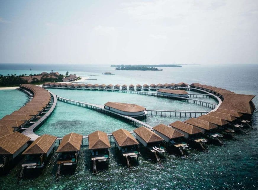 Vue aérienne d'un complexe hôtelier avec des bungalows sur pilotis en arc de cercle sur l'océan turquoise.