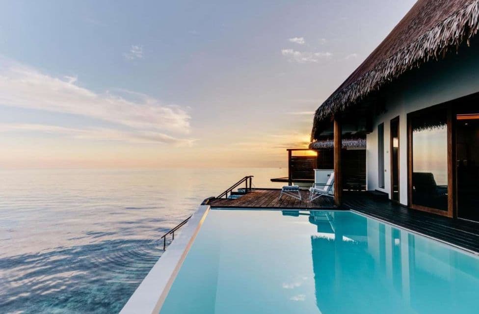 Un bungalow sull'acqua con tetto di paglia e piscina a sfioro si affaccia sull'oceano al tramonto.