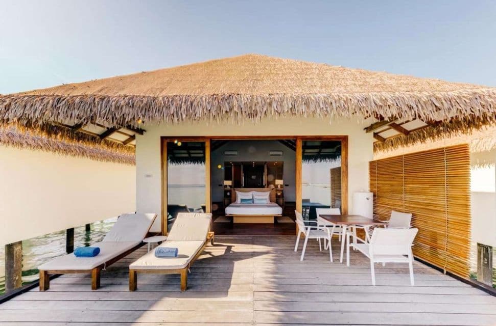 Un bungalow sull'acqua con tetto in paglia presenta un ponte in legno con sedie a sdraio e un tavolo, con la camera da letto visibile all'interno.