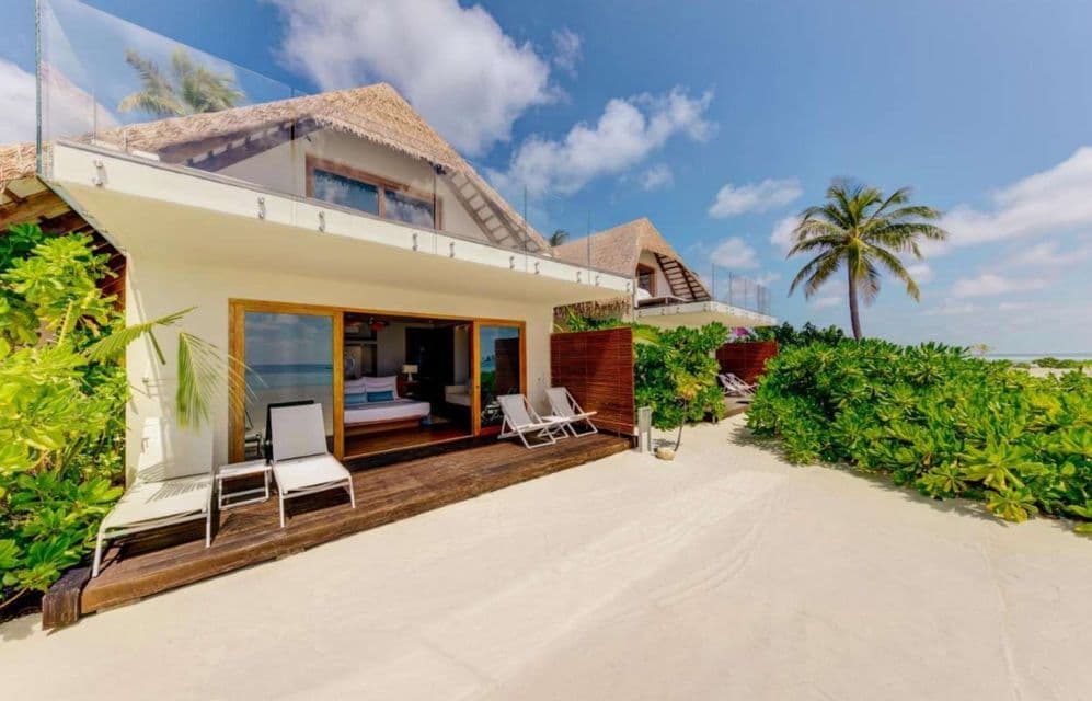 Una villa fronte mare con tetto di paglia e una terrazza in legno con sedie a sdraio, situata su una spiaggia di sabbia bianca accanto a piante tropicali.