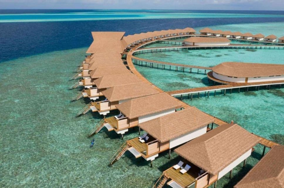 Une vue aérienne de bungalows sur pilotis aux toits de chaume reliés par des passerelles au-dessus d'une mer turquoise.