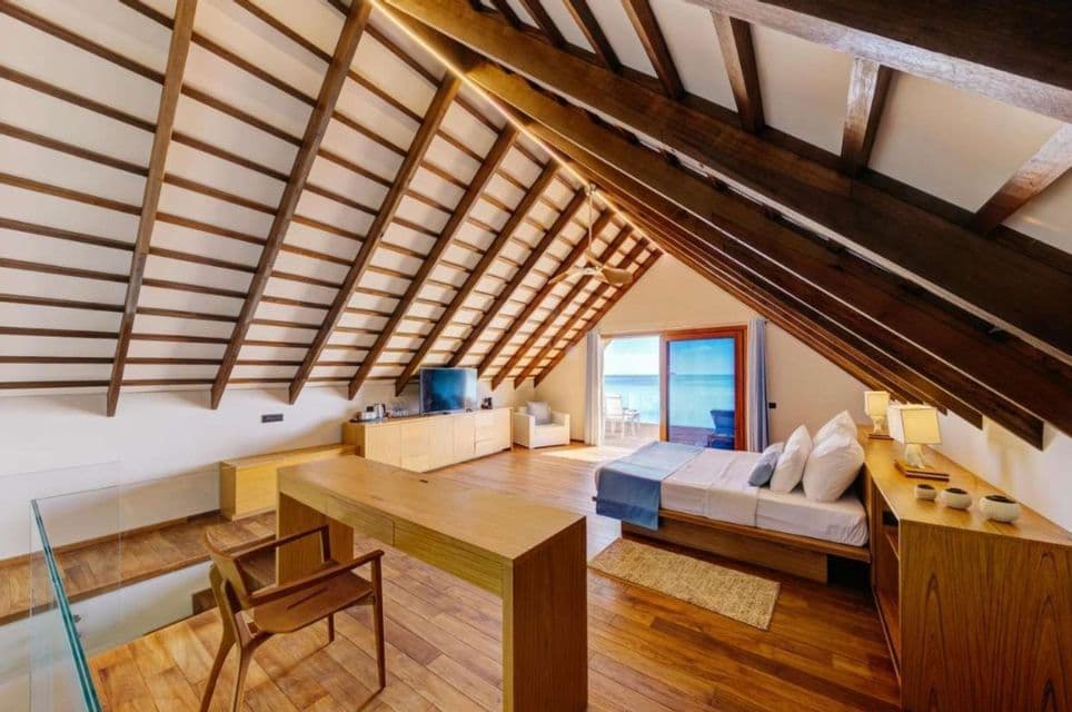 Ein luftiges Loft-Schlafzimmer mit Holzböden und offener Balkendecke, das sich zu einem Balkon mit Blick auf das türkisfarbene Meer öffnet.