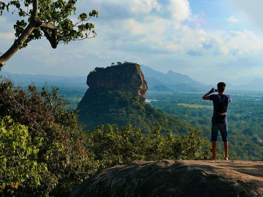 Desde un punto elevado, una persona fotografía una gran formación rocosa rodeada de un denso bosque verde y montañas.