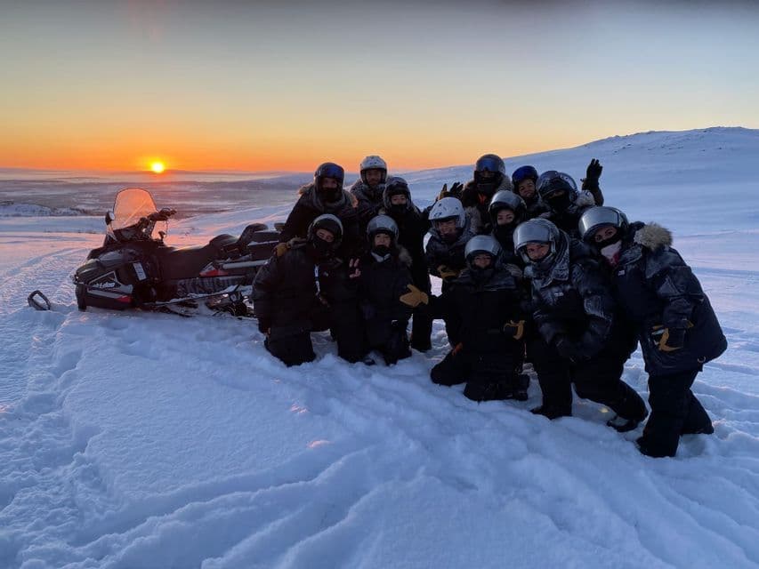Un gruppo WeRoad in posa per una foto in un paesaggio innevato accanto a una motoslitta al tramonto.