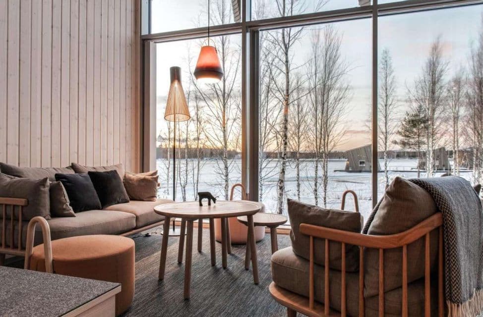 Un soggiorno moderno, rivestito in legno, con un divano, si affaccia attraverso una grande finestra su un paesaggio innevato con un lago ghiacciato.