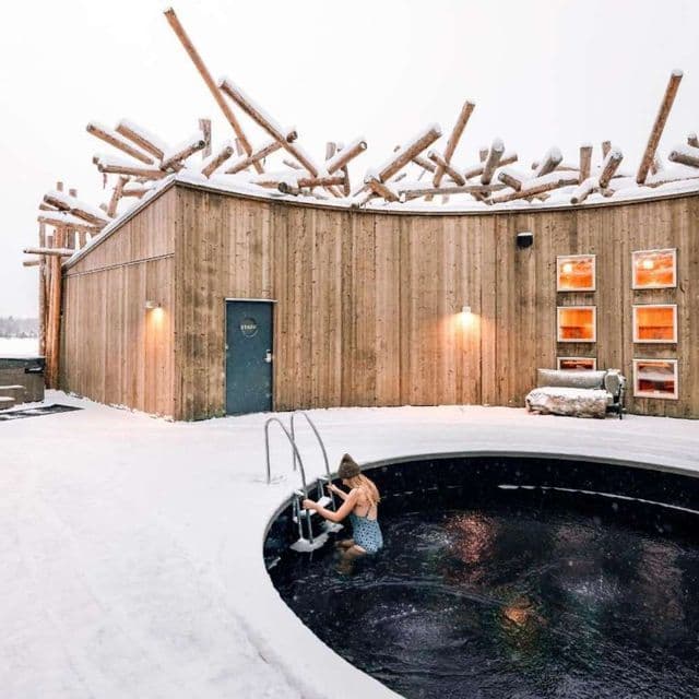 Una donna in costume e berretto sale una scaletta in una piscina esterna circolare e buia, circondata dalla neve, accanto a un edificio di legno.